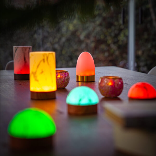 Op een houten tafel staan decoratieve lampen, waaronder de Tafellamp DORSETTO oplaadbaar 105 uur-waterdicht, marmeren groene houtlook, met afstandsbediening en kandelaars die groen, rood, oranje en roze oplichten. De onscherpe achtergrond creëert een gezellige buitensfeer.